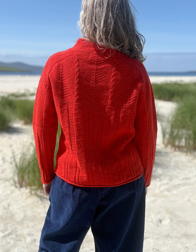 A person standing outdoors on a sandy beach wearing a bright red cable-knit sweater and navy pants, viewed from behind.