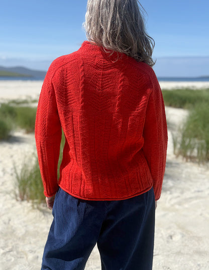 A person standing outdoors on a sandy beach wearing a bright red cable-knit sweater and navy pants, viewed from behind.