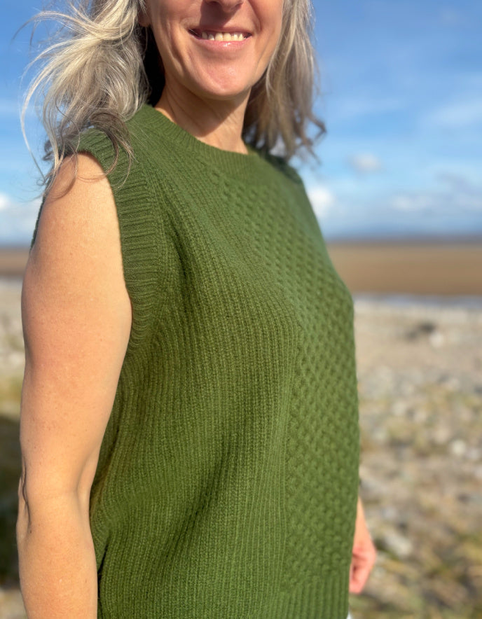 Woman wearing a green sleeveless knitted top standing on a beach with a blue sky.