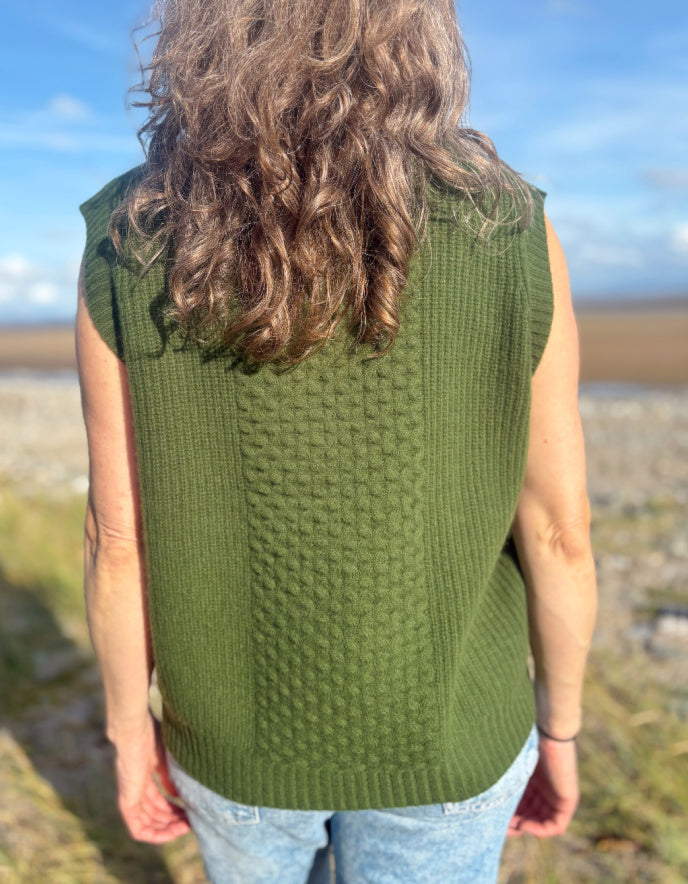 Person wearing a green knitted vest standing outdoors with a scenic background