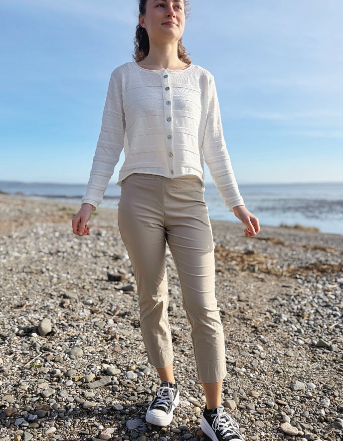 Woman standing on a pebbly beach wearing a white cardigan and beige pants.