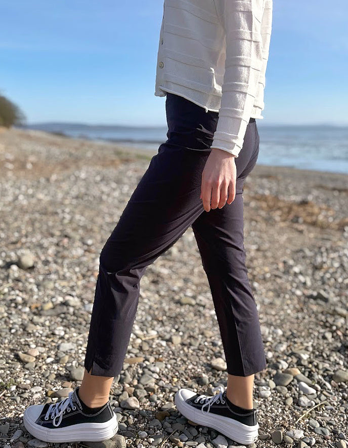 Person wearing black pants and white sneakers on a pebbly beach with ocean in the background