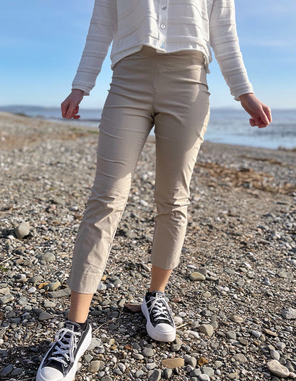 Person wearing beige pants and black sneakers standing on a pebbly beach with ocean in the background