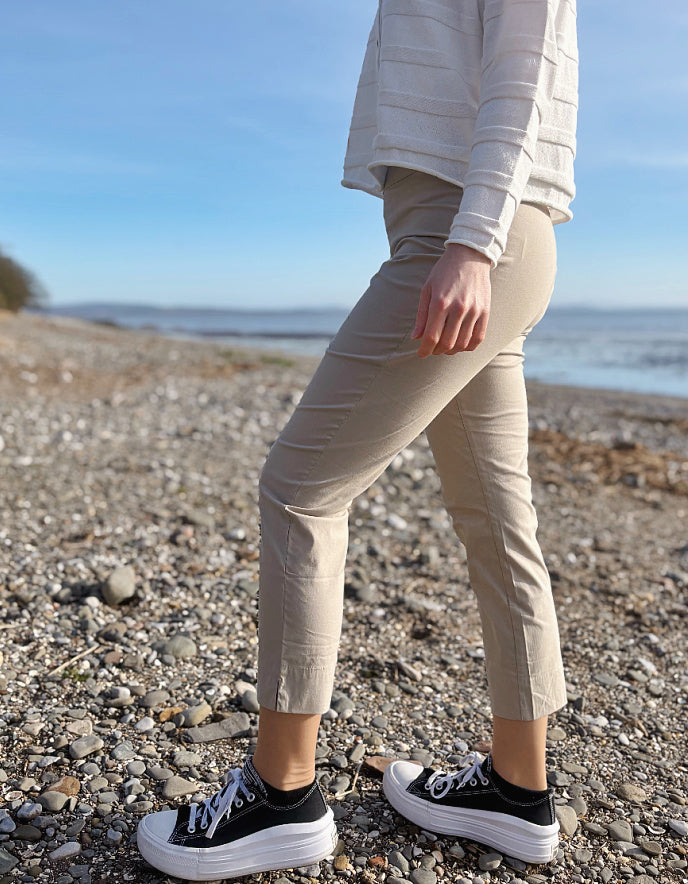 Person wearing beige pants and black sneakers on a pebbly beach with ocean in the background