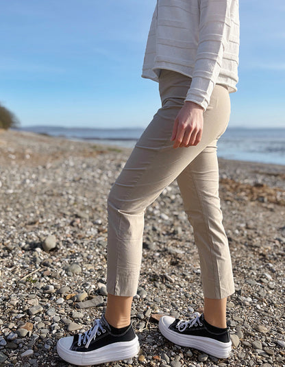 Person wearing beige pants and black sneakers on a pebbly beach with ocean in the background