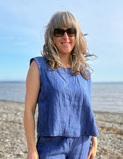 Woman in a blue sleeveless top and pants standing on a beach with sunglasses.