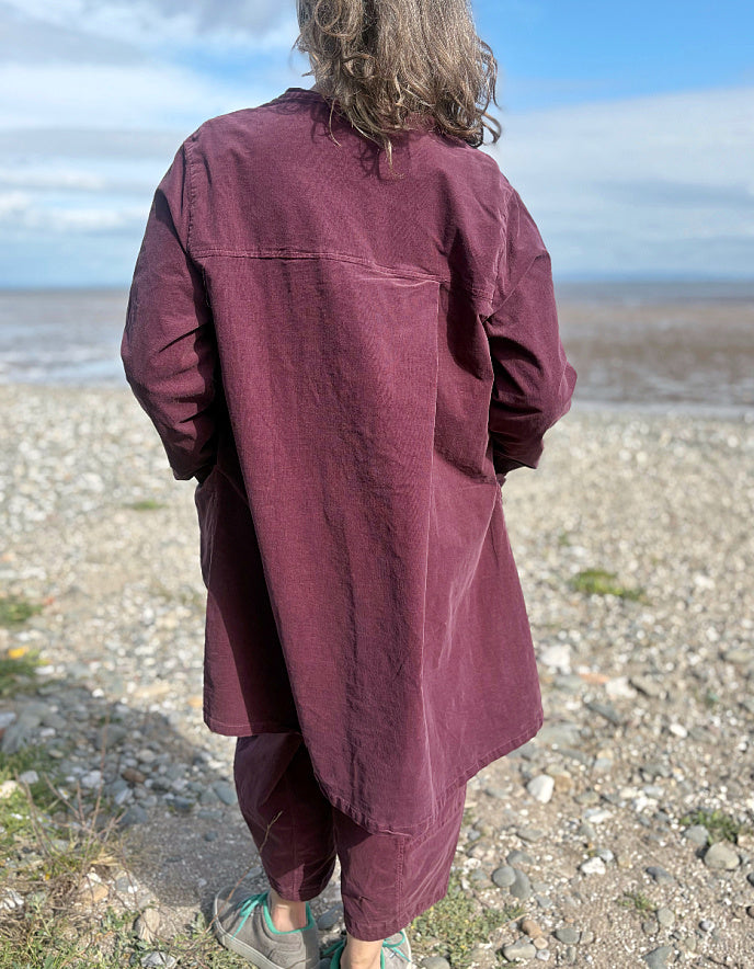 Woman in a burgundy plum corduroy duster coat and lantern shaped trousers standing on a pebbly beach with ocean in the background