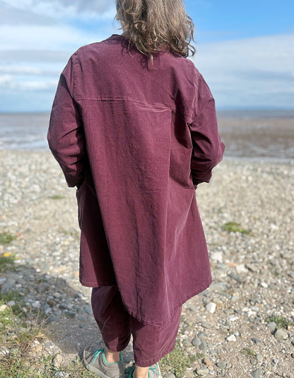 Woman in a burgundy plum corduroy duster coat and lantern shaped trousers standing on a pebbly beach with ocean in the background