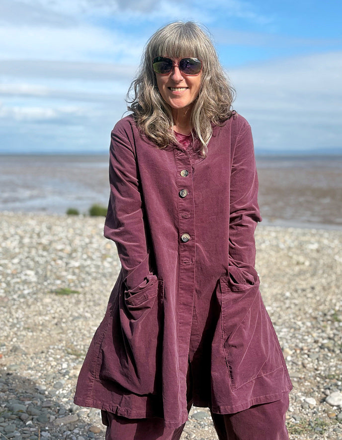 Woman in a burgundy plum corduroy duster coat and lantern shaped trousers standing on a pebbly beach with ocean in the background