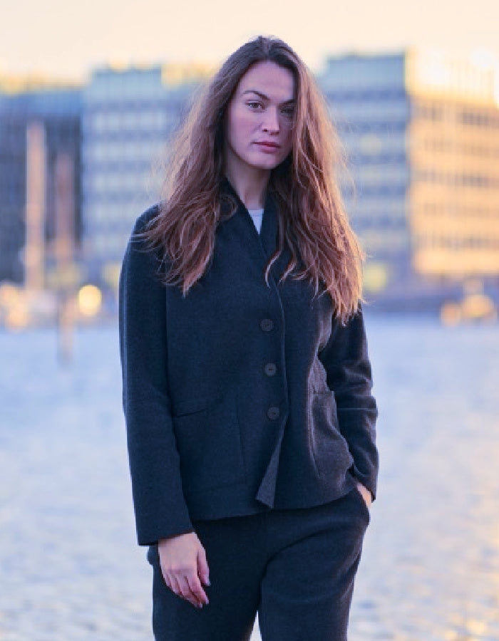 A woman with long light brown hair stands outdoors wearing a dark navy tailored jacket and matching dark trousers, with a softly blurred city waterfront in the background.