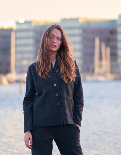 A woman with long light brown hair stands outdoors wearing a dark navy tailored jacket and matching dark trousers, with a softly blurred city waterfront in the background.