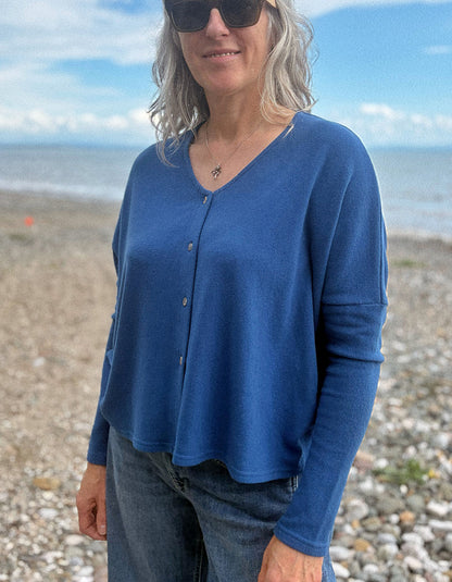 A woman wearing a blue long-sleeve button-front top and jeans, standing on a pebble beach with the ocean and sky in the background.