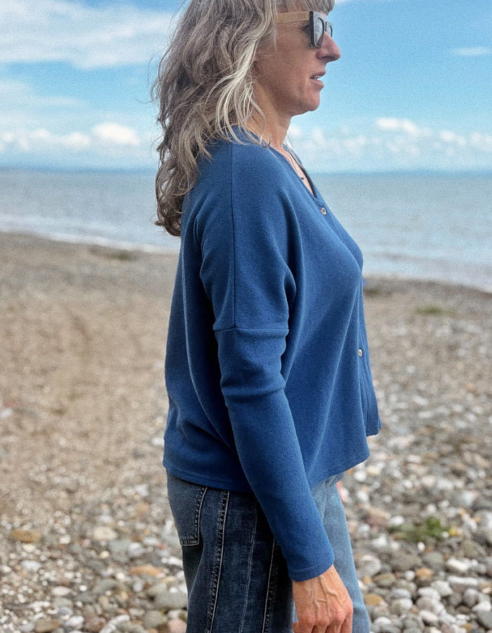 A woman wearing a blue long-sleeve knit top and jeans stands in profile on a pebble beach with the ocean and sky in the background.