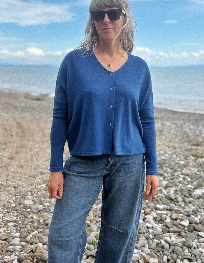 A woman wearing a blue long-sleeve button-front top and blue jeans stands on a pebble beach with the sea and sky in the background.