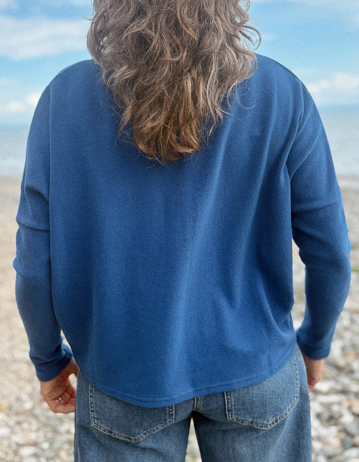 A person viewed from behind wearing a blue long-sleeve top and blue jeans, standing outdoors on a pebble beach with water and sky in the background.
