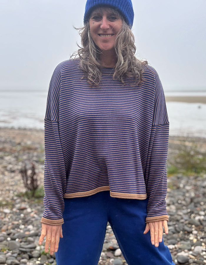 A woman wearing a blue beanie and a blue and camel striped knit sweater, standing on a pebble beach with water in the background.