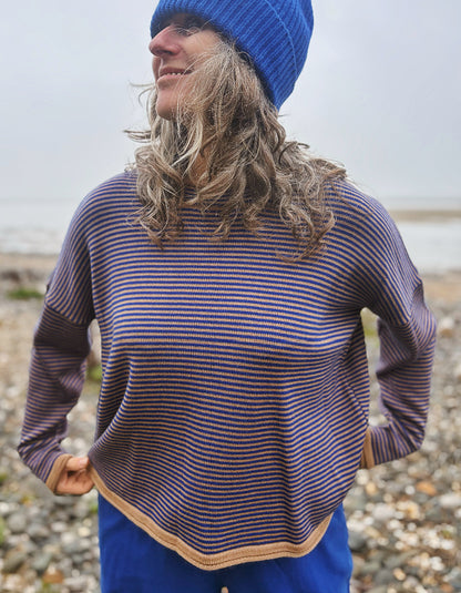 A woman wearing a blue beanie and a blue and camel striped knit sweater, standing on a pebble beach with water in the background.