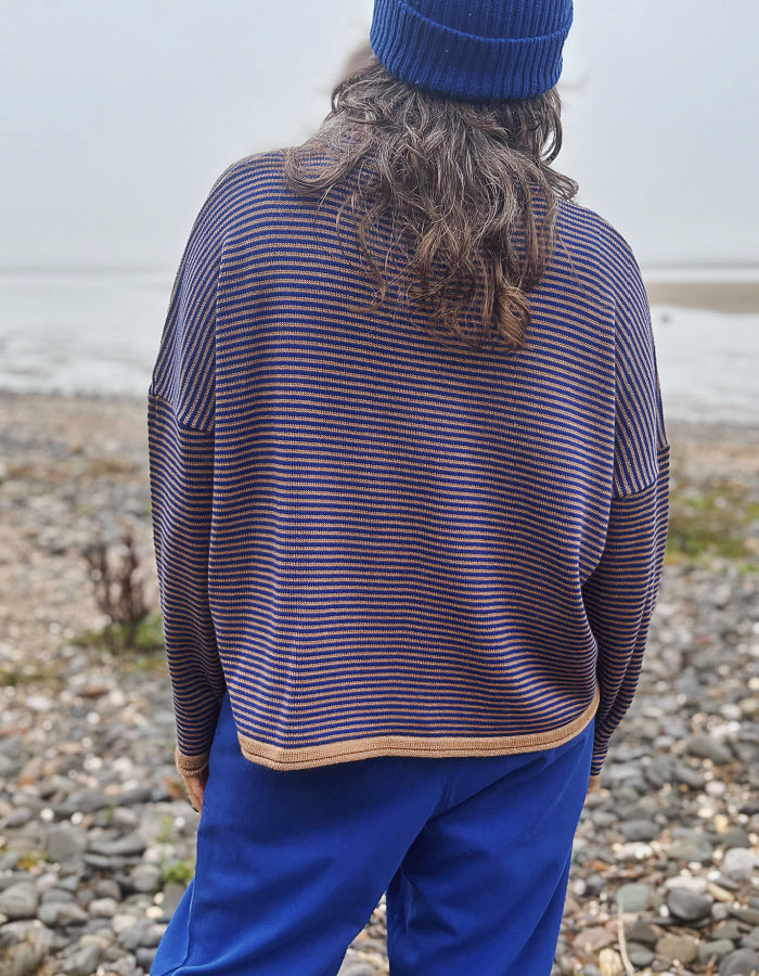 A woman wearing a blue beanie and a blue and camel striped knit sweater, standing on a pebble beach with water in the background.