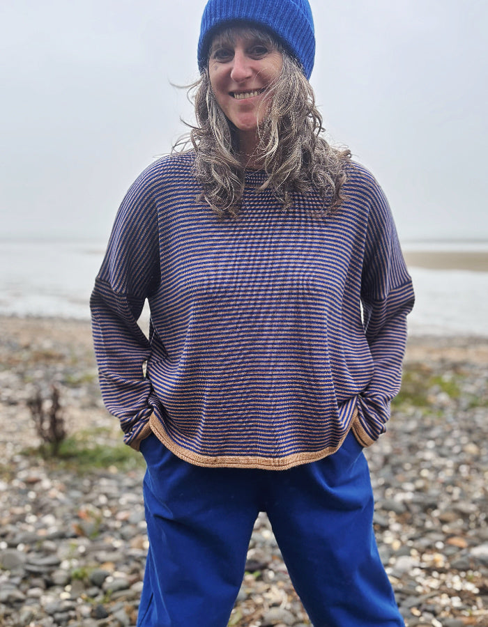 A woman wearing a blue beanie and a blue and camel striped knit sweater, standing on a pebble beach with water in the background.
