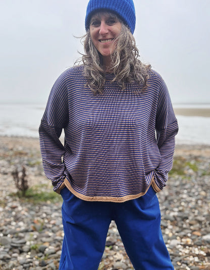 A woman wearing a blue beanie and a blue and camel striped knit sweater, standing on a pebble beach with water in the background.
