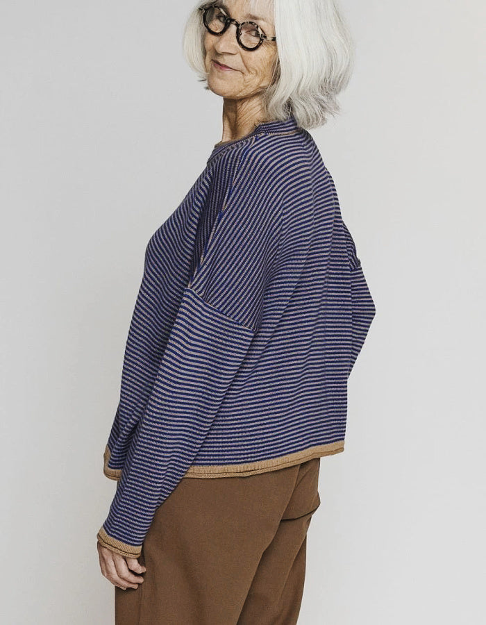 An older woman with short white hair and glasses is shown from the side, wearing a navy and blue striped long-sleeve top with brown pants, against a plain light gray background.