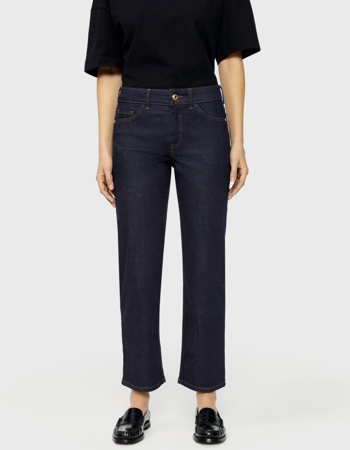 A model wearing dark navy blue cropped jeans with a straight-leg fit, paired with a black t-shirt and black leather loafers, shot against a light gray studio background.