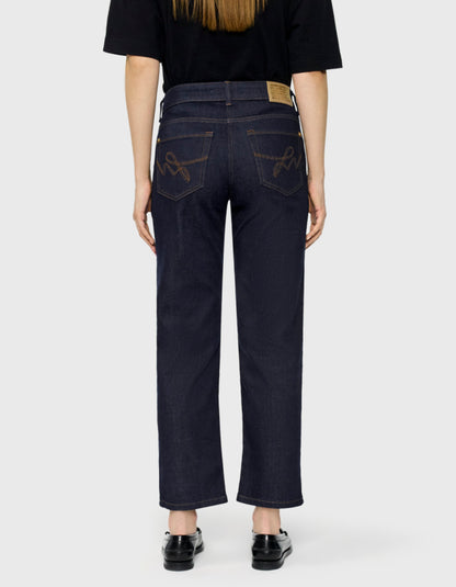 Back view of a person wearing dark indigo denim jeans with decorative brown embroidery on the back pockets, paired with a black t-shirt and black shoes, against a light gray background.