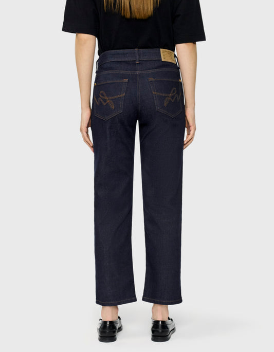 Back view of a person wearing dark indigo denim jeans with decorative brown embroidery on the back pockets, paired with a black t-shirt and black shoes, against a light gray background.