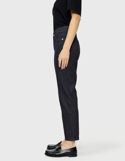 A side profile view of a person wearing dark indigo slim-fit jeans, a black t-shirt, and black leather loafers against a light gray studio background.