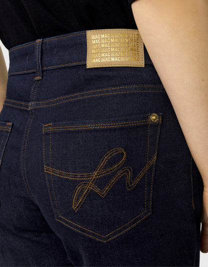 Close-up of the back waistband and pocket area of dark indigo jeans on a model, showing a tan leather patch with repeating 'MAC' text and a gold embroidered script on the back pocket.