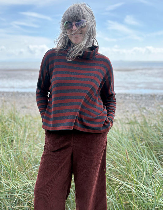 Person wearing a copper and charcoal striped turtle neck sweater with copper corduroy wide leg trousers and sunglasses on a beach
