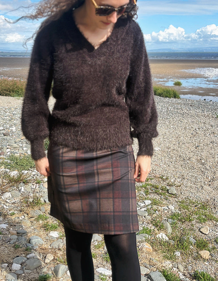 A person standing on a rocky beach wearing a dark brown fuzzy knit sweater, a brown and red plaid skirt, black tights, and sunglasses, with a body of water and cloudy sky in the background.