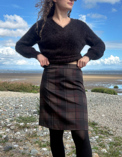 A woman stands outdoors on a rocky shoreline under a blue sky with clouds, wearing a dark fuzzy cropped sweater and a dark plaid skirt with black tights.