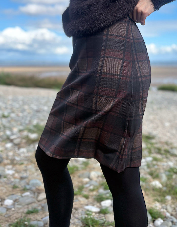 A person outdoors wearing a knee-length plaid skirt in dark brown, burgundy, and black tones, paired with black tights and a dark fuzzy top. The background shows a rocky shoreline with water and a cloudy sky.