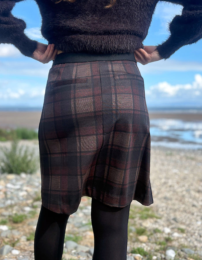 A person standing outdoors on a rocky shoreline wearing a dark plaid skirt with black tights and a fuzzy dark sweater, photographed from the back.