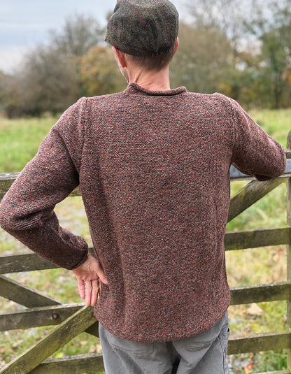 Man wearing a brown knitted sweater and cap standing by a wooden gate in a field.