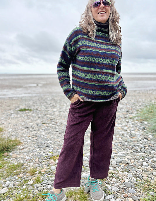 Person wearing a patterned sweater and purple pants standing on a pebbly beach.