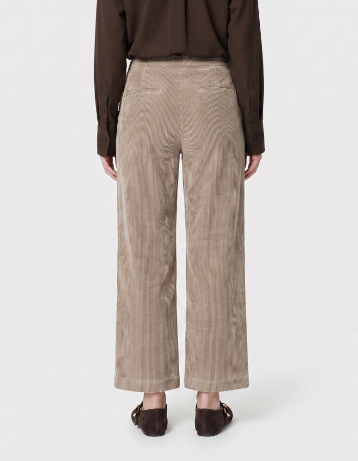 A person wearing wide-leg cropped trousers in a taupe corduroy fabric, paired with white sneakers and a white top, shown from the waist down on a light grey studio background.