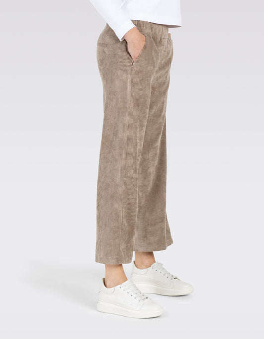 A person wearing wide-leg cropped trousers in a taupe corduroy fabric, paired with white sneakers and a white top, shown from the waist down on a light gray studio background.