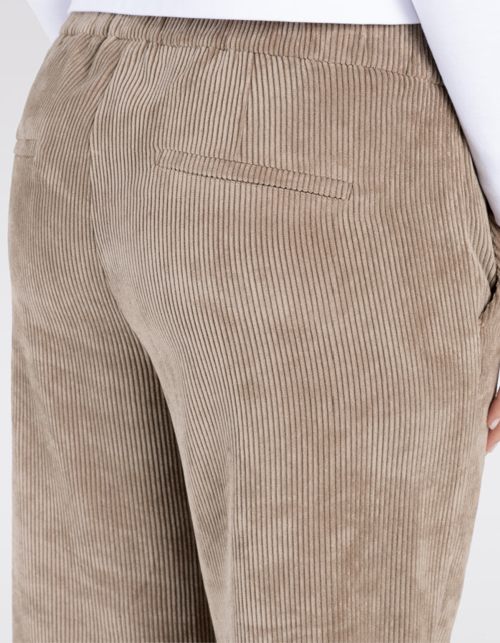 A person wearing wide-leg cropped trousers in a taupe corduroy fabric, paired with white sneakers and a white top, shown from the waist down on a light grey studio background.