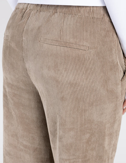 A person wearing wide-leg cropped trousers in a taupe corduroy fabric, paired with white sneakers and a white top, shown from the waist down on a light grey studio background.