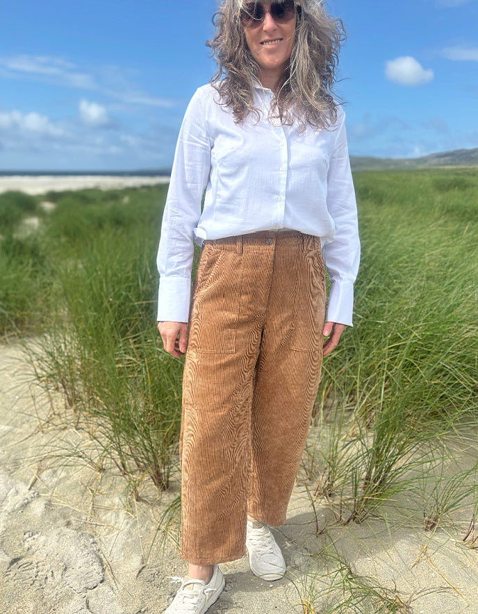 A woman standing on a sandy beach dune wearing a white long-sleeve button-up shirt, brown corduroy wide-leg pants, and white sneakers, with grass, sky, and ocean in the background.