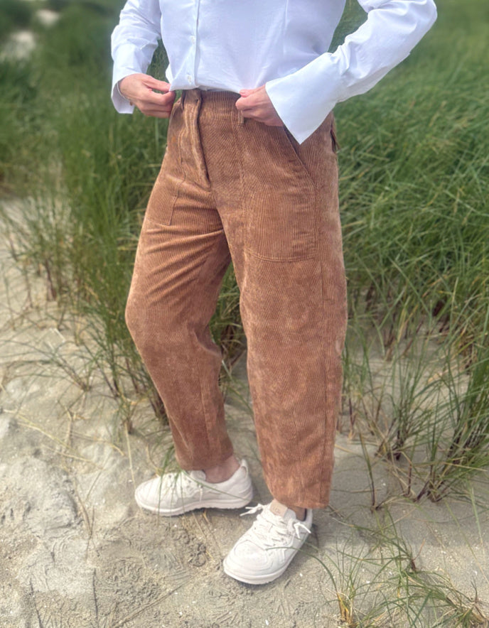 A person standing outdoors on sand dunes wearing brown corduroy pants, a white button-up shirt, and white sneakers.