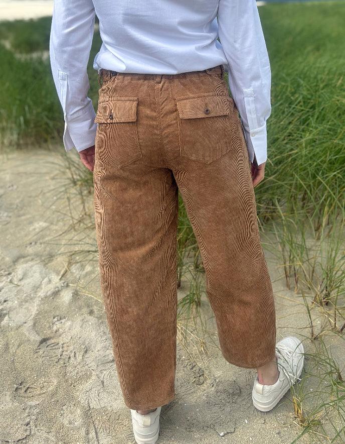 A person viewed from behind wearing brown corduroy pants, a white long-sleeve shirt, and white sneakers, standing on a sandy path with green grass in the background.