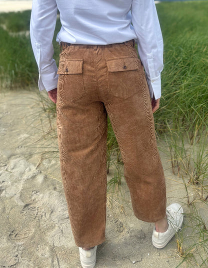 A person viewed from behind wearing brown corduroy pants, a white long-sleeve shirt, and white sneakers, standing on a sandy path with green grass in the background.