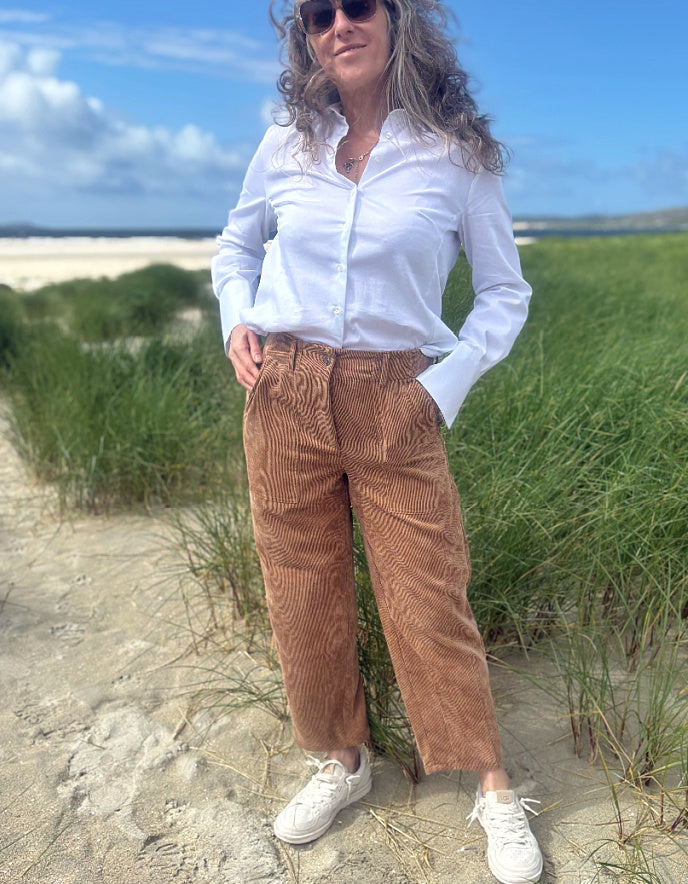A woman standing on a sandy beach dune wearing brown corduroy trousers, a white button-up shirt, and white sneakers, with green grass and blue sky in the background.