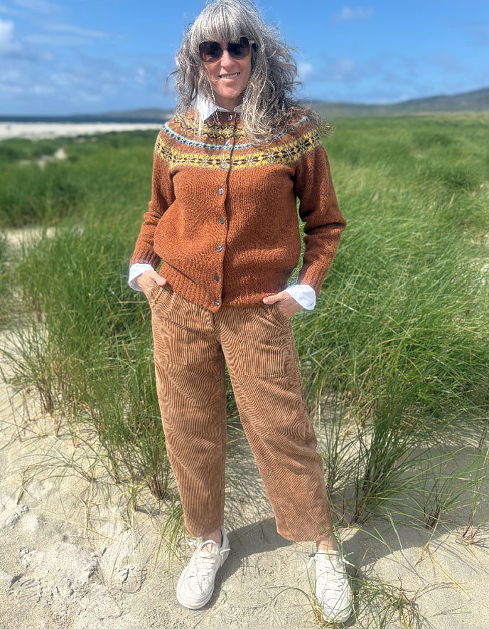 A woman standing on sandy dunes wearing a rust-brown button-up cardigan with a multicolor patterned yoke, brown corduroy pants, a white collared shirt, and white sneakers, with tall beach grass and a blue sky in the background.