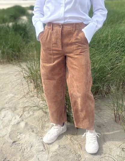 A person standing on sand near grass, wearing brown corduroy wide-leg pants, a white button-up shirt, and white sneakers.