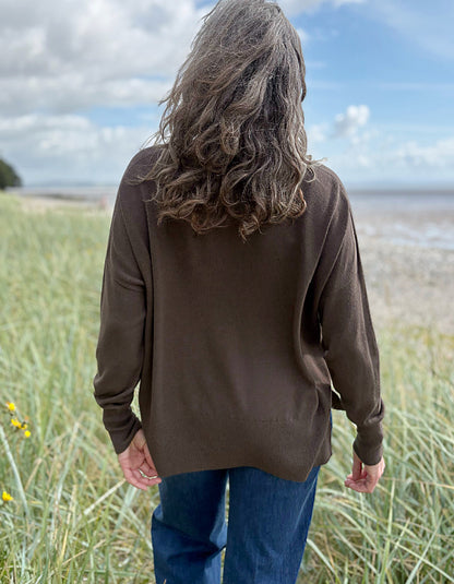 Person wearing a brown sweater with mock turtle neck standing in grass with a beach and blue sky in the background