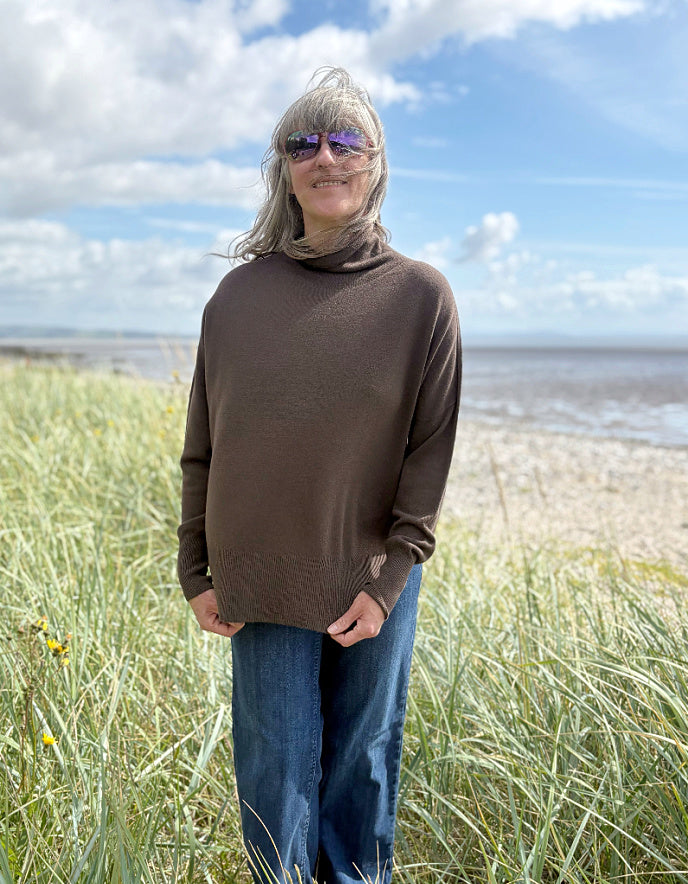 Person wearing a brown sweater with mock turtle neck standing in grass with a beach and blue sky in the background